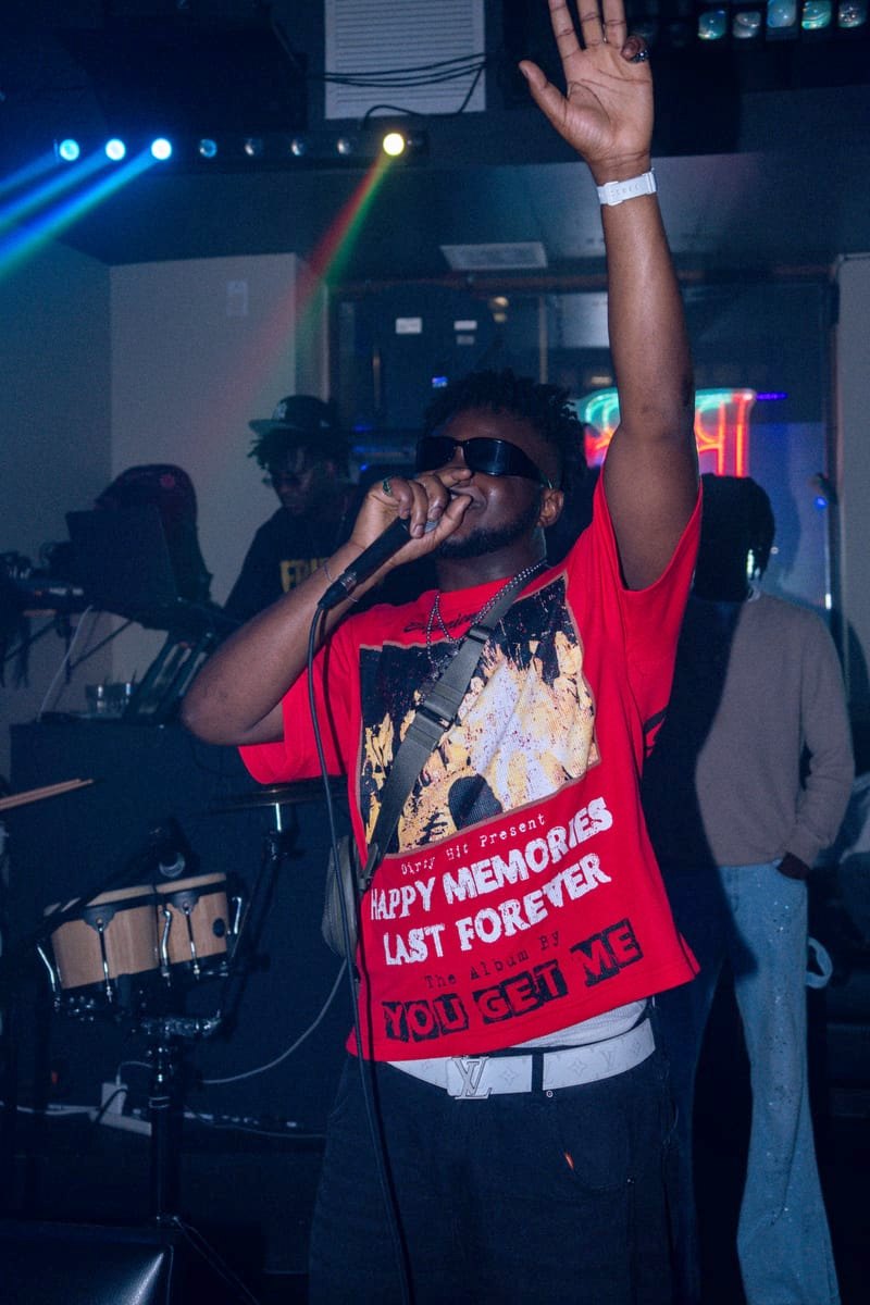Performer in a red graphic tee raising one hand and singing into the mic under the stage lights