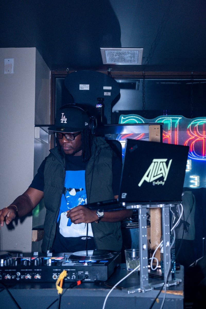 DJ in a green vest and cap working the decks behind a laptop