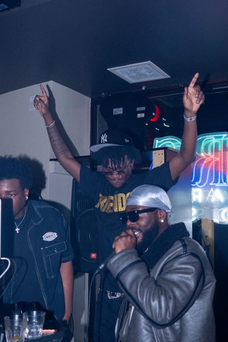 Artist on the mic in a leather jacket while a friend celebrates with both arms raised behind him at the DJ booth