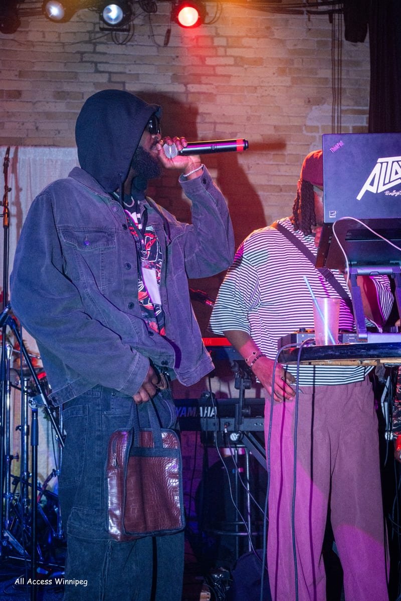 Hooded performer on the mic beside the DJ setup, warm stage light
